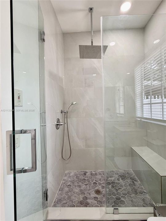 Walk-in shower featuring a frameless glass enclosure, ceiling-mounted rainfall showerhead, handheld sprayer, built-in bench, and grey hexagonal floor tile