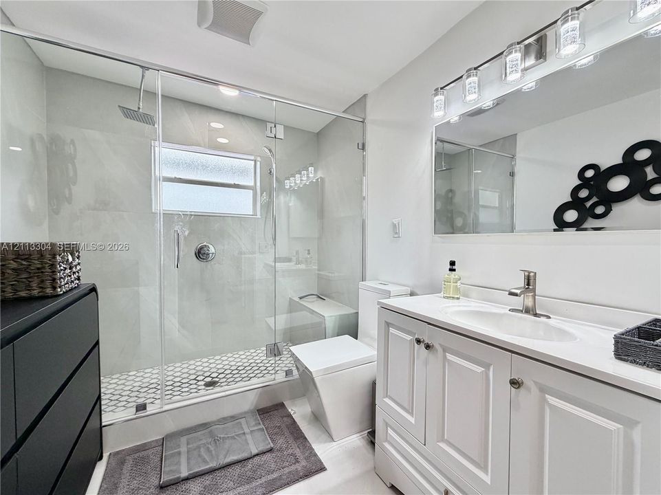 Bathroom featuring a frameless glass-enclosed shower with large-format wall tile and hexagonal floor tile, a white vanity cabinet with an integrated sink, and a multi-bulb light fixture above a wide mirror