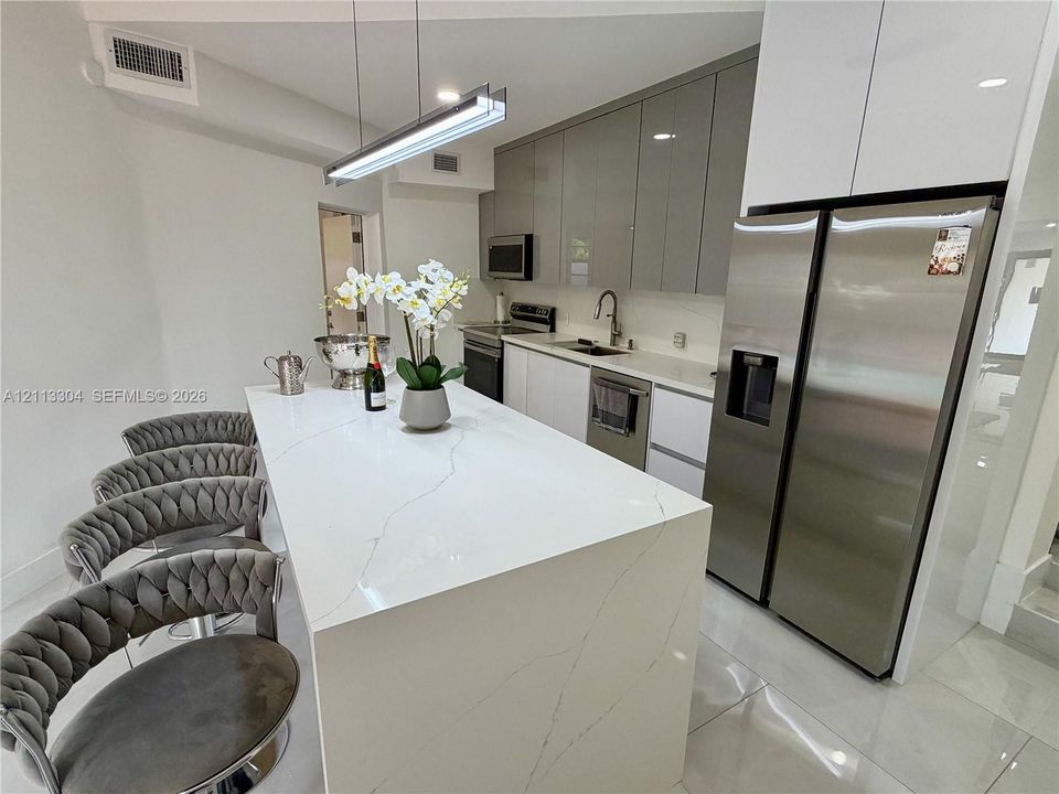 Kitchen featuring a waterfall-edge island with stone-finish countertop, high-gloss cabinetry, stainless steel appliances, and linear pendant lighting