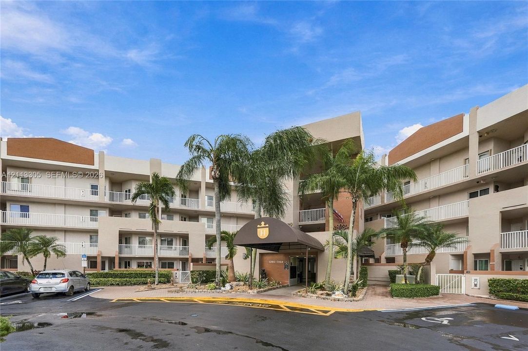 Multi-story residential complex featuring a covered porte-cochere entrance, recessed balconies with white railings, and manicured landscaping with mature palm trees