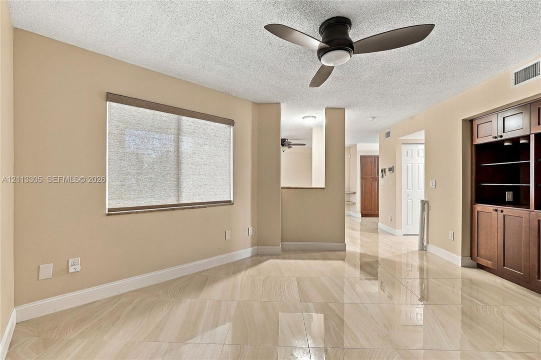 Polished stone-finish tile flooring, built-in dark wood cabinetry with open shelving, matte black three-blade ceiling fan, and window featuring a roller shade
