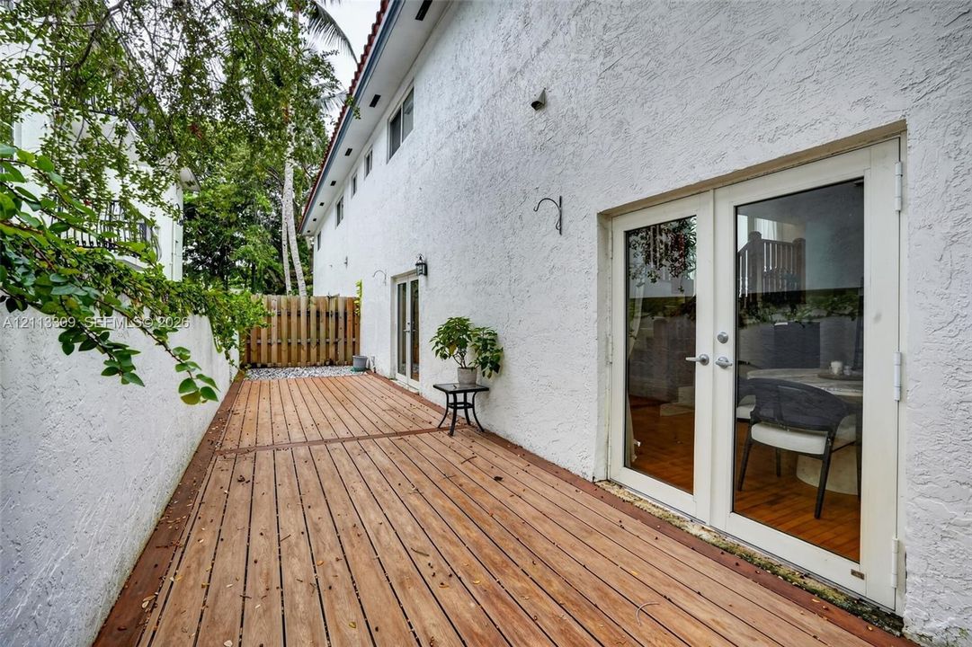Exterior wood-finish decking featuring dual glass-paned double doors, white textured stucco walls, and a perimeter wood fence