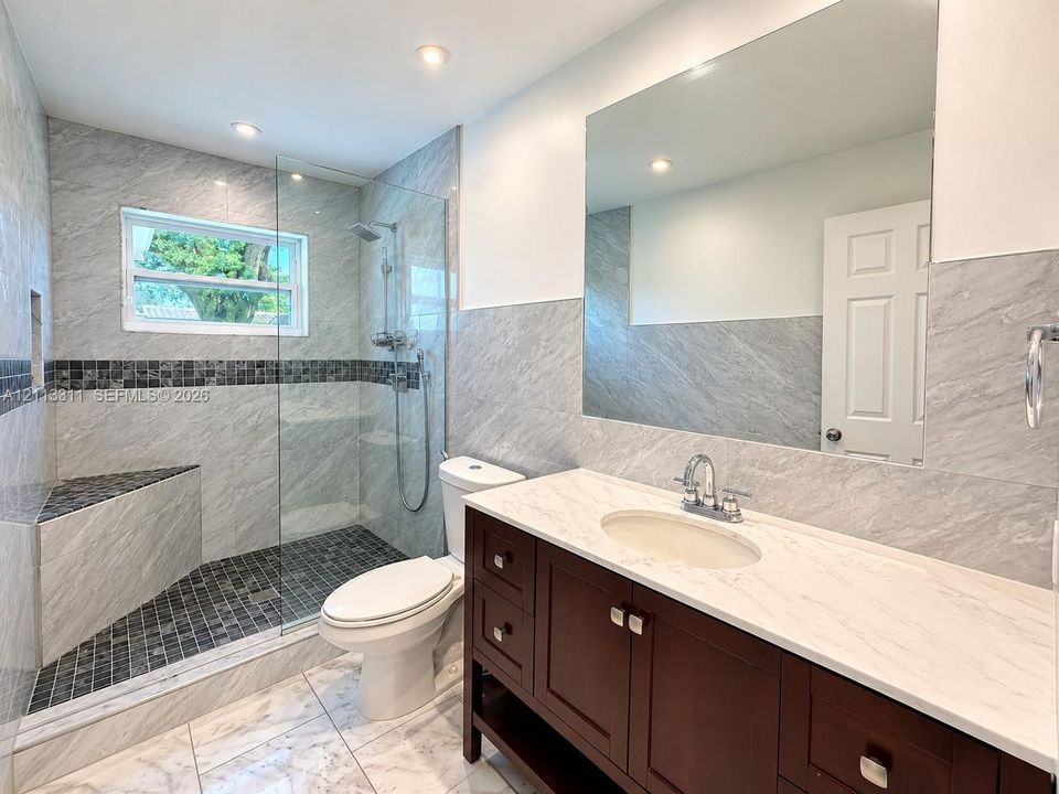 Bathroom featuring a wood-finish vanity with a light-toned countertop, a walk-in shower with a glass panel and built-in bench, recessed lighting, and large-format wall tiling with a mosaic accent border