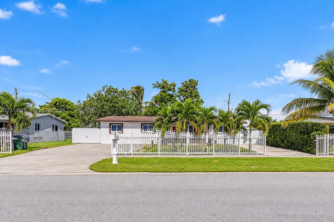 Single-story residence featuring a white exterior, expansive concrete driveway, full-property white metal fencing, and mature palm trees