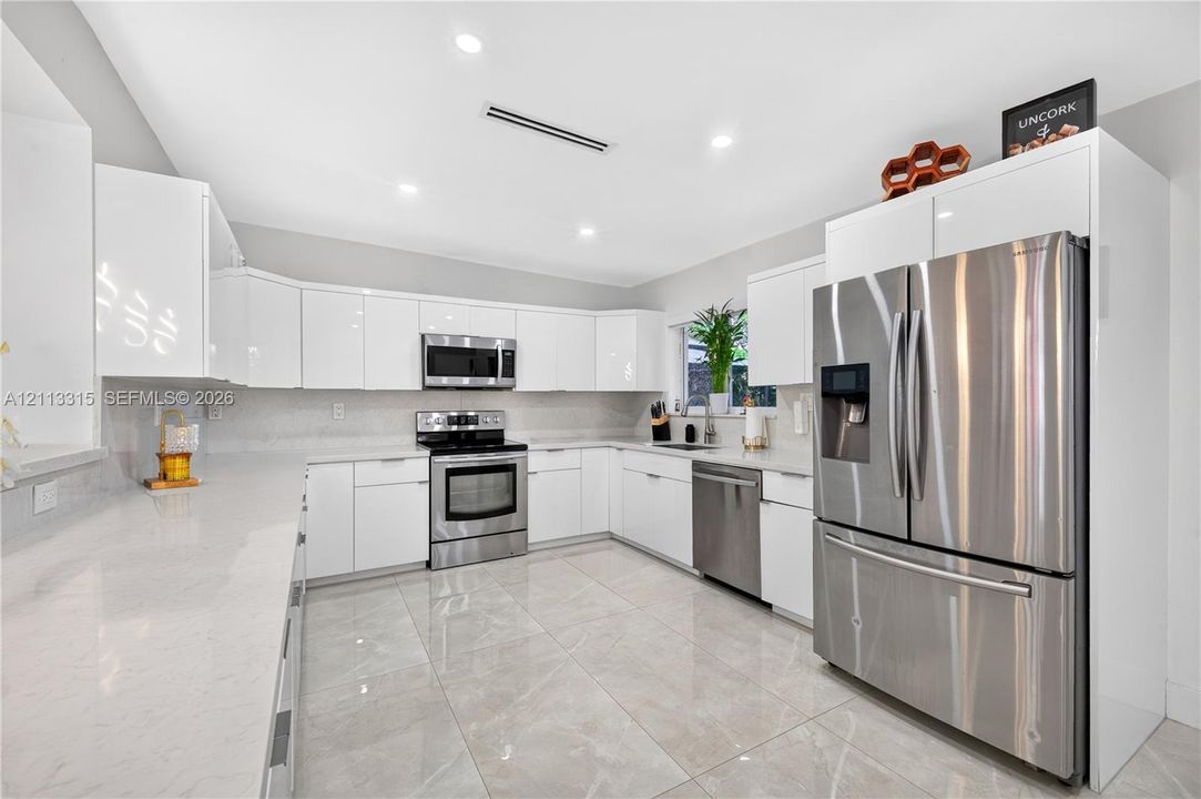 Kitchen featuring high-gloss white cabinetry, light stone-finish countertops, stainless steel appliances, and large-format tile flooring