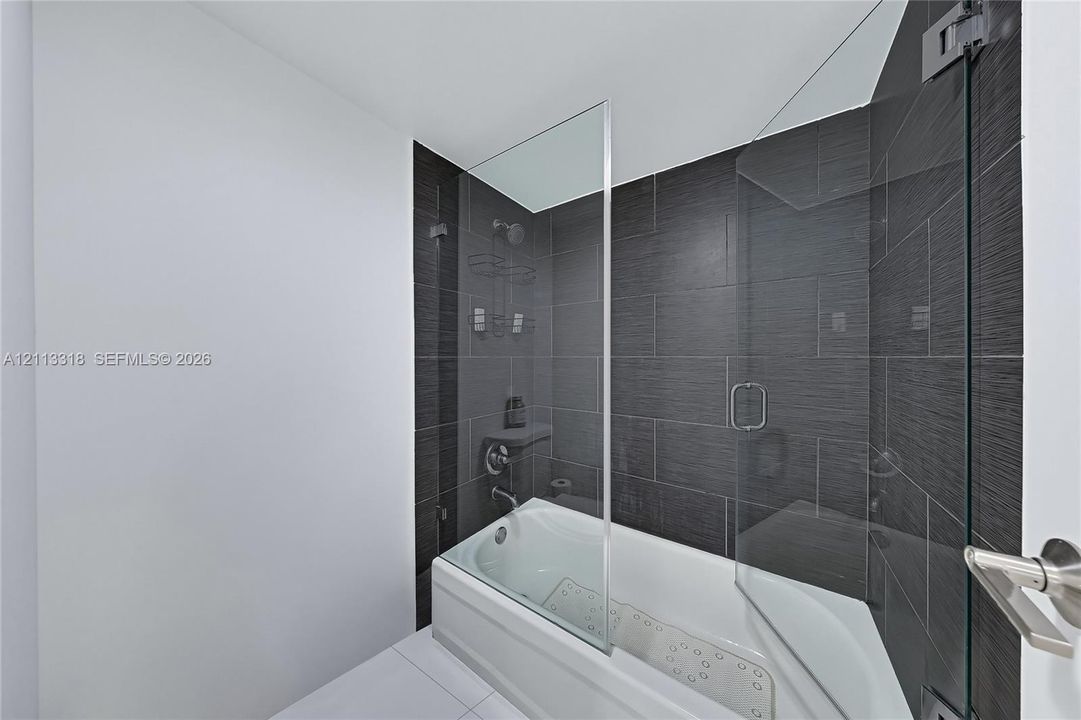Bathtub with a glass panel, dark charcoal textured wall tile, chrome shower fixtures, and minimalist hardware