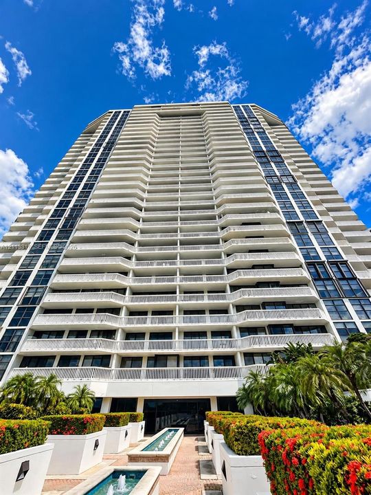 High-rise residential exterior featuring stacked concrete balconies, floor-to-ceiling glass windows, manicured perimeter landscaping, and stone-tiled fountain features