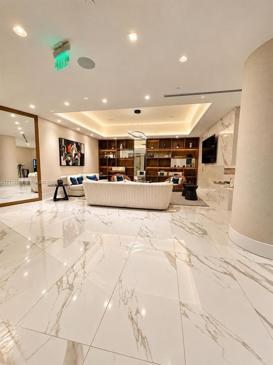 Large-format polished stone flooring, recessed tray ceiling with integrated cove lighting, floor-to-ceiling wooden built-in shelving, full-height wall mirror with metallic frame, and stone-surround fireplace feature