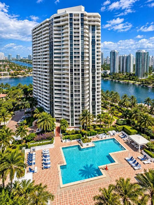 High-rise residential tower featuring wrap-around balconies, a ground-level swimming pool, brick-paver pool deck, covered poolside pavilion, and lush tropical landscaping