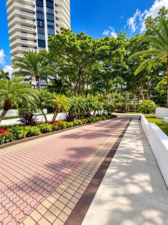 Paved walkway featuring interlocking brick patterns, concrete borders, mature palm trees, landscaping, and tall structural architecture