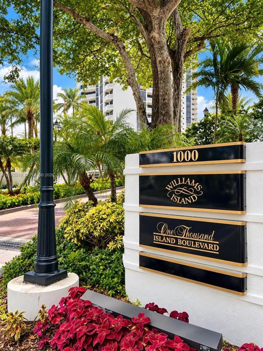 Monument signage featuring gold-lettered panels on a white stucco base, surrounded by mature palm trees, professional landscaping, and a brick-paved entry drive
