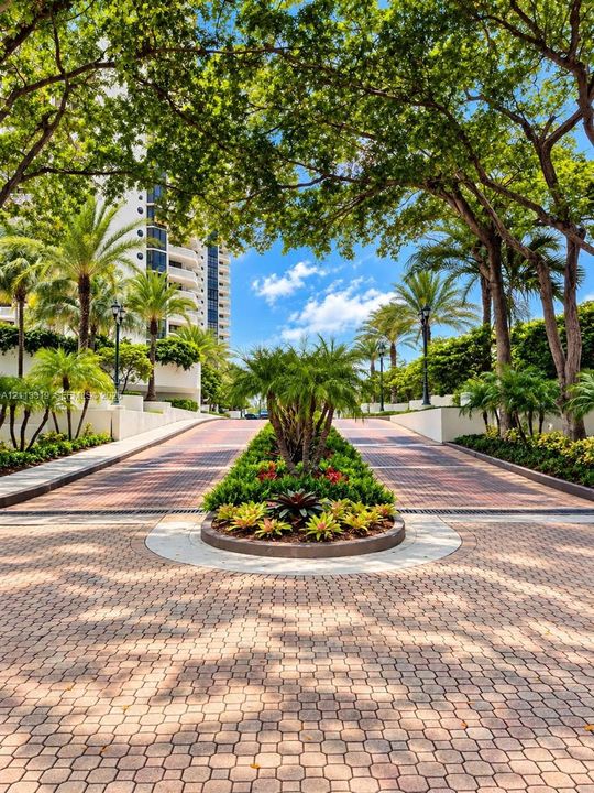 Paved circular drive featuring a central garden bed, mature canopy trees, palm landscaping, and decorative brick-style pavers