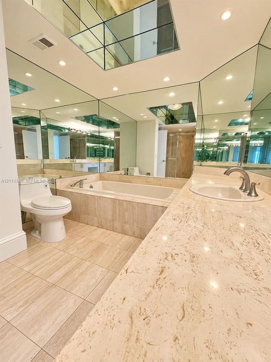 Stone-surfaced vanity with an integrated basin, inset soaking tub, tile flooring, and wall-to-ceiling mirror panels