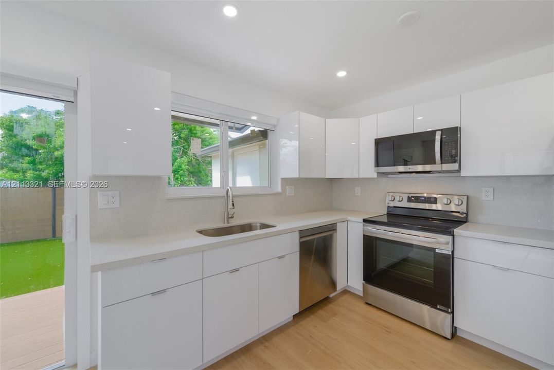 Kitchen featuring high-gloss white cabinetry, stainless steel appliances, light-toned stone countertops, recessed lighting, and wood-finish flooring
