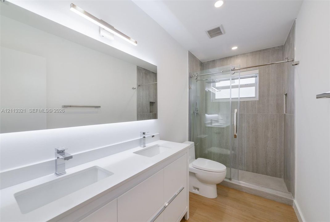 Dual-sink vanity with white stone-finish countertop, under-mount sinks, chrome faucets, full-width mirror with integrated lighting, and glass-enclosed shower with floor-to-ceiling tile