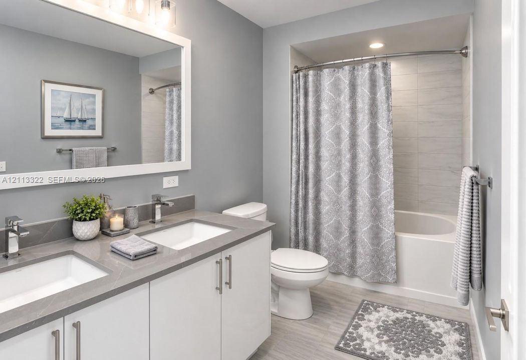 Bathroom featuring a dual-sink vanity with white cabinetry and solid surface countertops, expansive framed mirror, and a tub-shower combination with light-toned horizontal wall tile