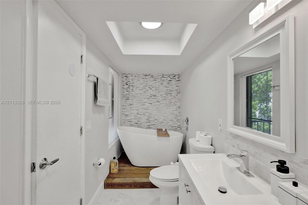 Freestanding soaking tub set upon a wood-finish platform, stone-tiled feature wall, recessed skylight, vanity with an integrated basin, and framed wall mirror
