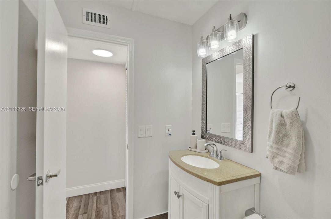White vanity with stone-composite countertop, oval undermount sink, metallic framed wall mirror, multi-bulb vanity light fixture, and wood-finish flooring