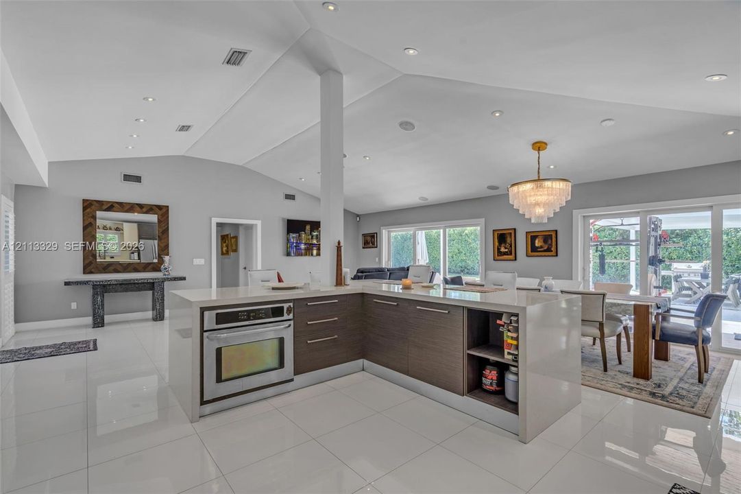 Open-concept interior featuring a vaulted ceiling, recessed lighting, polished large-format tile flooring, and a kitchen island with built-in oven and wood-finish cabinetry