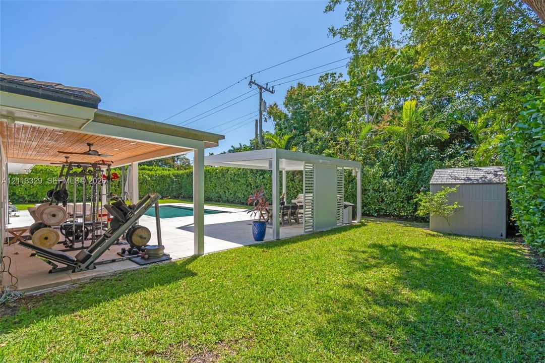 Backyard featuring a swimming pool, a covered patio with a beadboard ceiling, a separate metal-framed pergola, and a storage shed surrounded by mature perimeter hedging