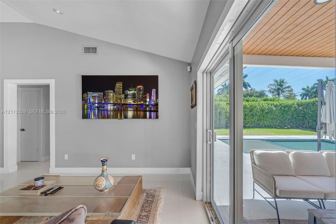Vaulted ceilings, large-format tile flooring, and floor-to-ceiling sliding glass doors opening to a covered patio with wood-paneled ceiling, manicured landscaping, and a pool