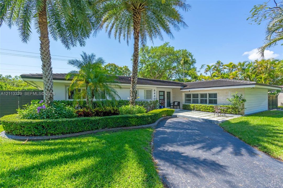 Single-story residence featuring a low-pitched roof, manicured hedges, mature palm trees, stone-finish exterior accents, and a paved front patio area