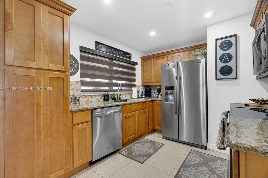 Kitchen featuring medium-toned wood cabinetry, stone countertops, tiled backsplash, stainless steel appliances, and large-format floor tile