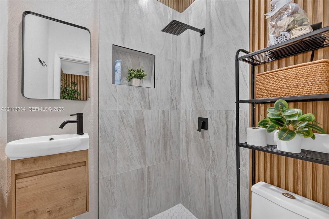 Wall-mounted wood-finish vanity with a rectangular sink and matte black faucet, wall-mounted mirror with black frame, walk-in shower featuring floor-to-ceiling stone-style tile, built-in shower niche, and wood-slat accent wall