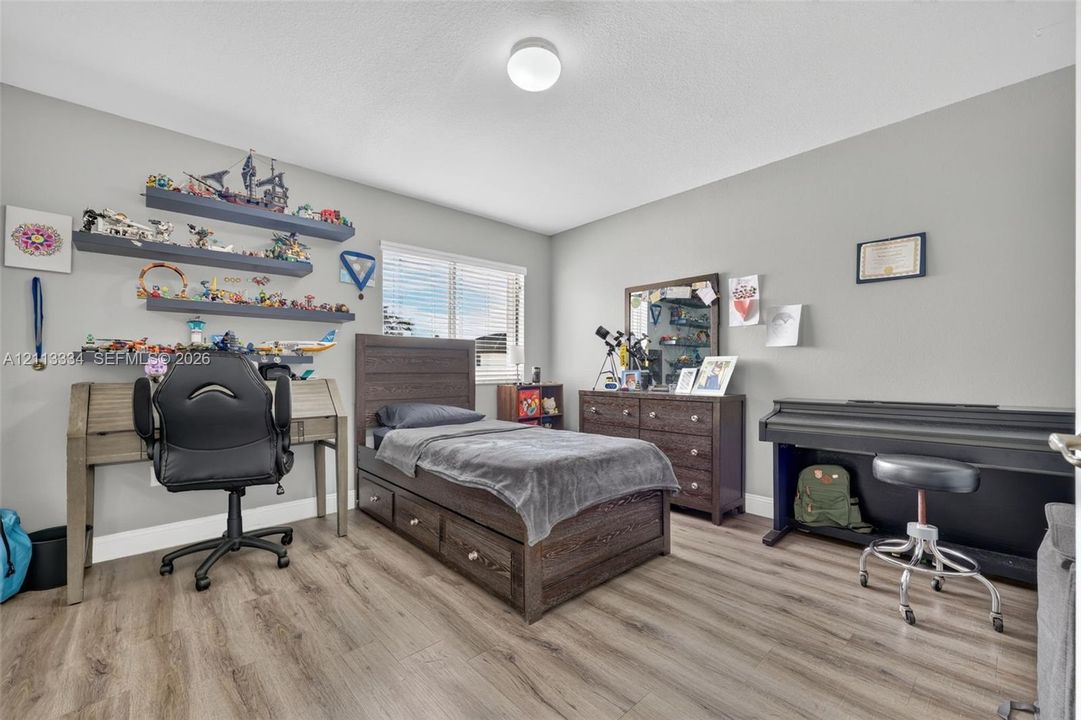 Neutral interior room featuring wood-finish flooring, three wall-mounted floating shelves, a single window with horizontal blinds, and a circular flush-mount ceiling light fixture