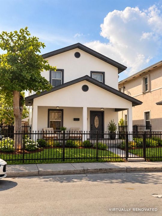 Two-story exterior featuring light-toned stucco facade, covered front porch with square columns, dark-trimmed windows, and decorative metal perimeter fencing