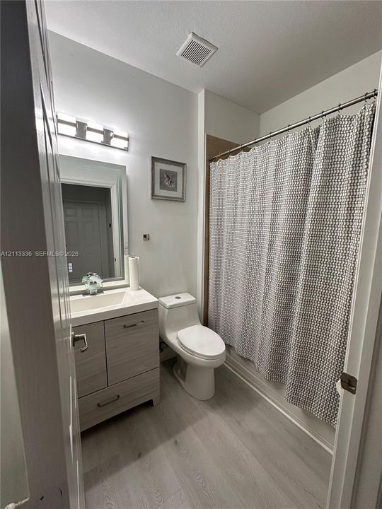 Bathroom featuring a wood-finish vanity with a rectangular sink, wall-mounted vanity lighting, a one-piece toilet, and wood-finish flooring