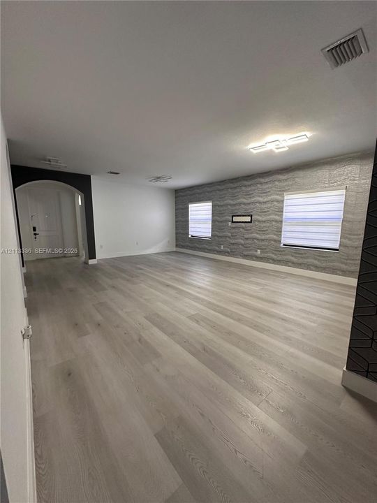 Open interior space featuring light-toned wood-finish flooring, a textured accent wall, integrated flush-mount ceiling lighting, and a prominent arched entryway
