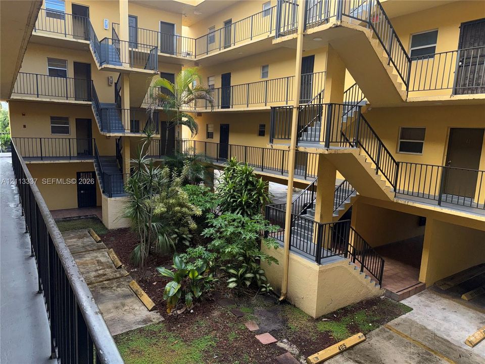 Multi-story building exterior featuring open-air corridors, metal staircases with black railings, and a central courtyard with established tropical landscaping