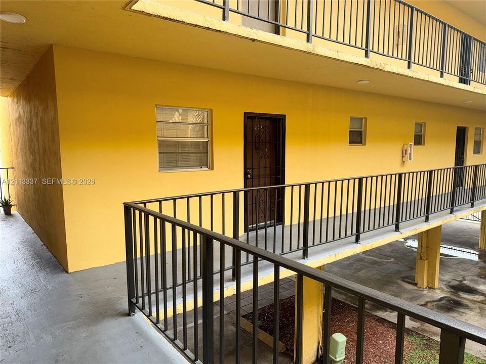 Exterior walkway featuring black metal railings and concrete flooring with access to building units