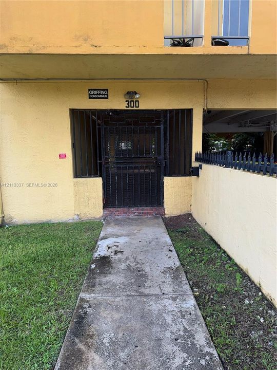 Concrete walkway leading to a gated entry with black iron security bars and exterior wall-mounted lighting