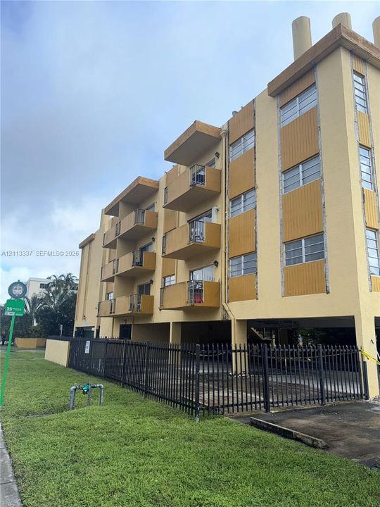 Multi-story building exterior featuring vertical panel siding, private cantilevered balconies, and a ground-level covered parking area enclosed by metal fencing