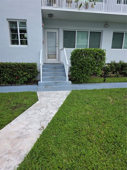 Exterior entrance featuring concrete stairs with white handrails, a glass-panel door, stone-paver walkway, and manicured hedges