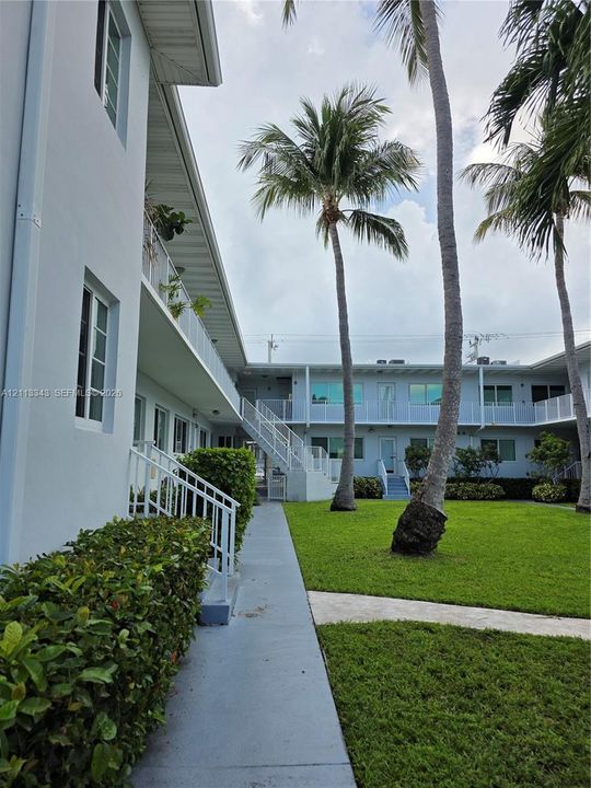 Multi-story exterior featuring light-colored stucco siding, metal staircases with white railings, manicured green space, and mature palm trees