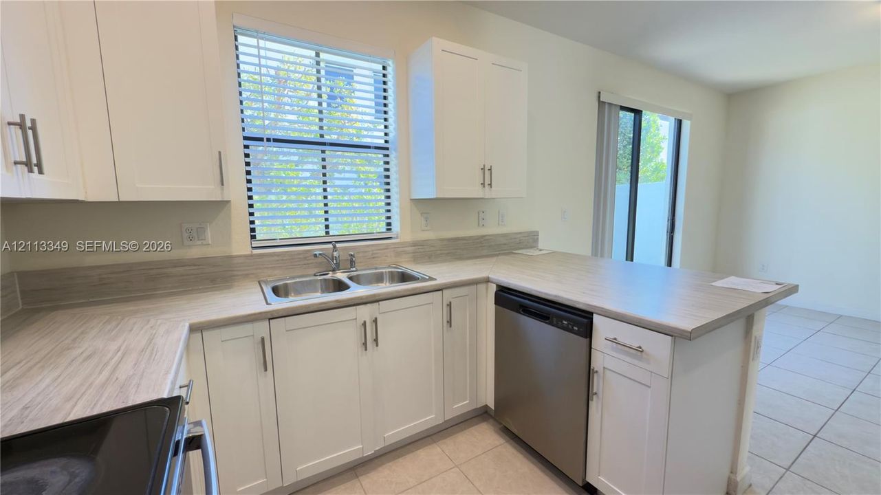 Kitchen featuring white shaker-style cabinetry, neutral countertops with integrated backsplash, stainless steel dishwasher, double-basin sink, and tile flooring