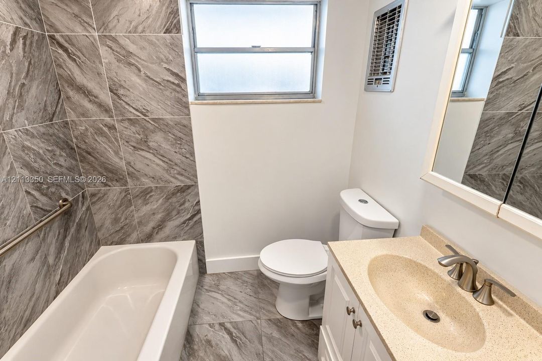 Bathroom featuring stone-finish wall and floor tiling, a white vanity with a composite countertop and integrated basin, a standard bathtub with a metallic grab bar, and a framed mirror