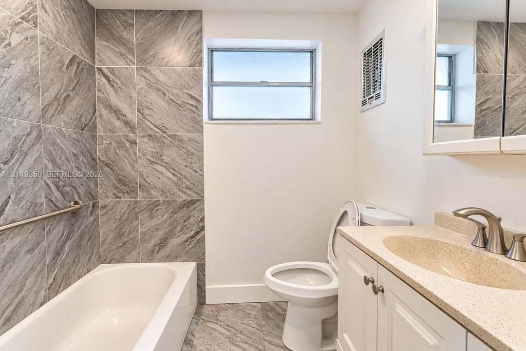 Bathroom featuring a tub-shower combination with large-format grey tile surround, stone-finish countertop, white cabinetry with metallic hardware, and a medicine cabinet with an integrated mirror