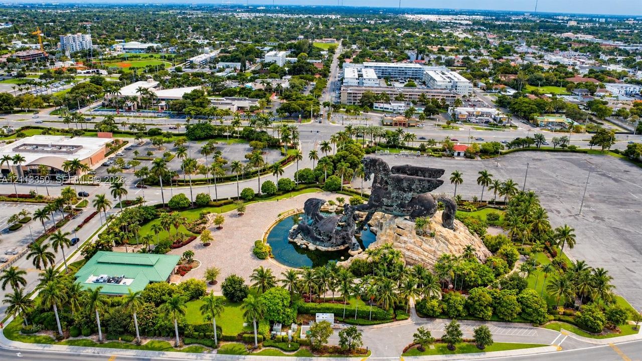 Aerial perspective featuring a large-scale sculptural monument, central water feature, paved courtyards, and extensive mature palm tree landscaping