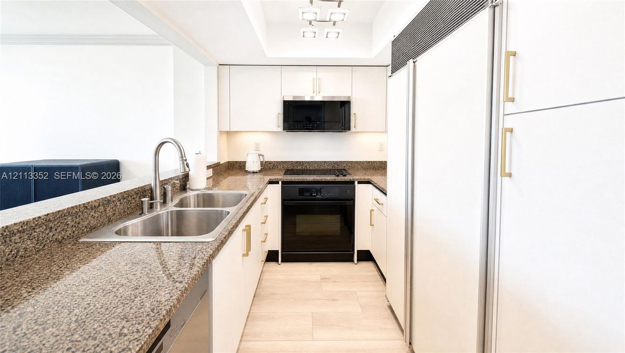 Kitchen featuring stone countertops, white flat-panel cabinetry with gold-tone hardware, stainless steel dual-basin sink with gooseneck faucet, built-in black appliances, and light-toned flooring