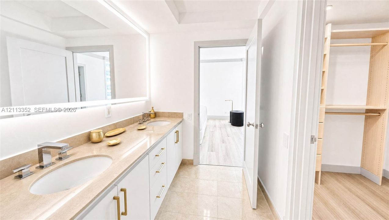 Dual-sink vanity with stone countertop, large illuminated mirror, white cabinetry featuring gold-tone hardware, and an open-concept walk-in closet with built-in shelving