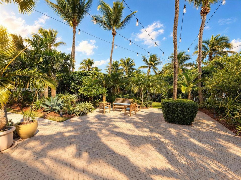 Outdoor patio featuring herringbone-patterned pavers, overhead string lighting, mature palm trees, dense perimeter greenery, and structural landscaping