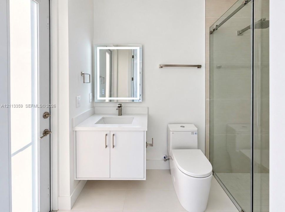 Floating vanity featuring a solid surface countertop, integrated sink, and brushed metal faucet, complemented by an illuminated rectangular mirror, a one-piece toilet, and a glass-enclosed shower with tile surround