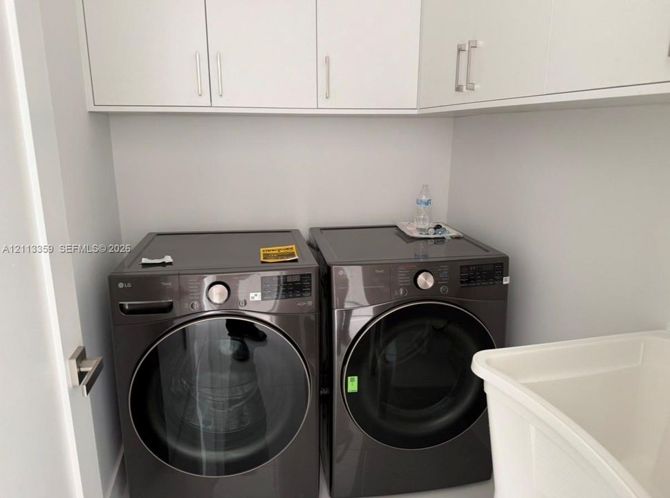 Laundry space featuring charcoal-finish front-load appliances, matte white shaker-style cabinetry with polished metal hardware, and a deep basin utility sink
