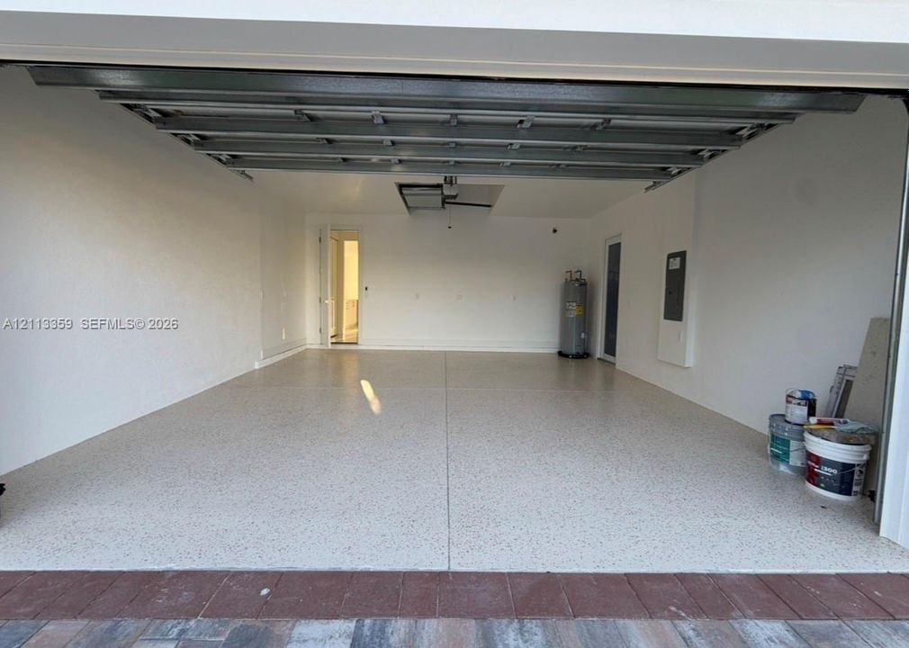 Finished garage featuring epoxy flooring, crisp white walls, exposed steel ceiling joists, an integrated water heater, and a recessed electrical panel