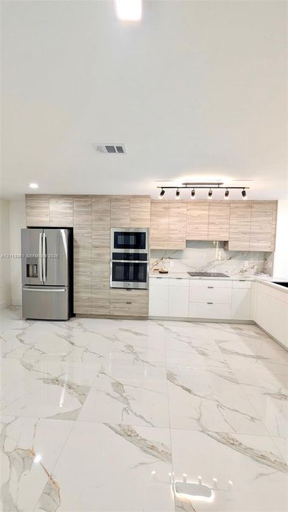 Kitchen featuring two-tone cabinetry with wood-finish upper cabinets, white lower cabinetry, integrated stainless steel appliances, stone-finish tile flooring, and multi-bulb track lighting
