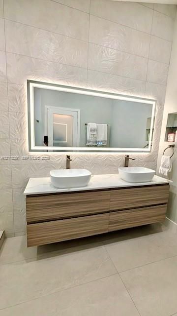 Floating wood-finish vanity with dual vessel sinks, quartz countertop, large backlit mirror, and textured wall tile